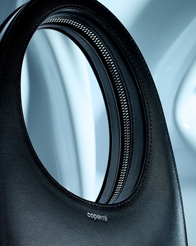 Close-up studio shot of a black leather Coperni handbag with a top handle and a silver zipper, set against a light blue background.