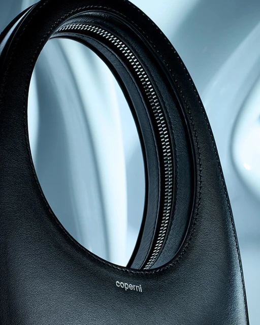 Close-up studio shot of a black leather Coperni handbag with a top handle and a silver zipper, set against a light blue background.