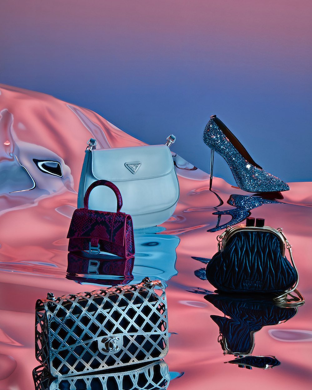Fashion still life featuring handbags and a high-heeled shoe on a reflective surface with a blue and pink gradient background.