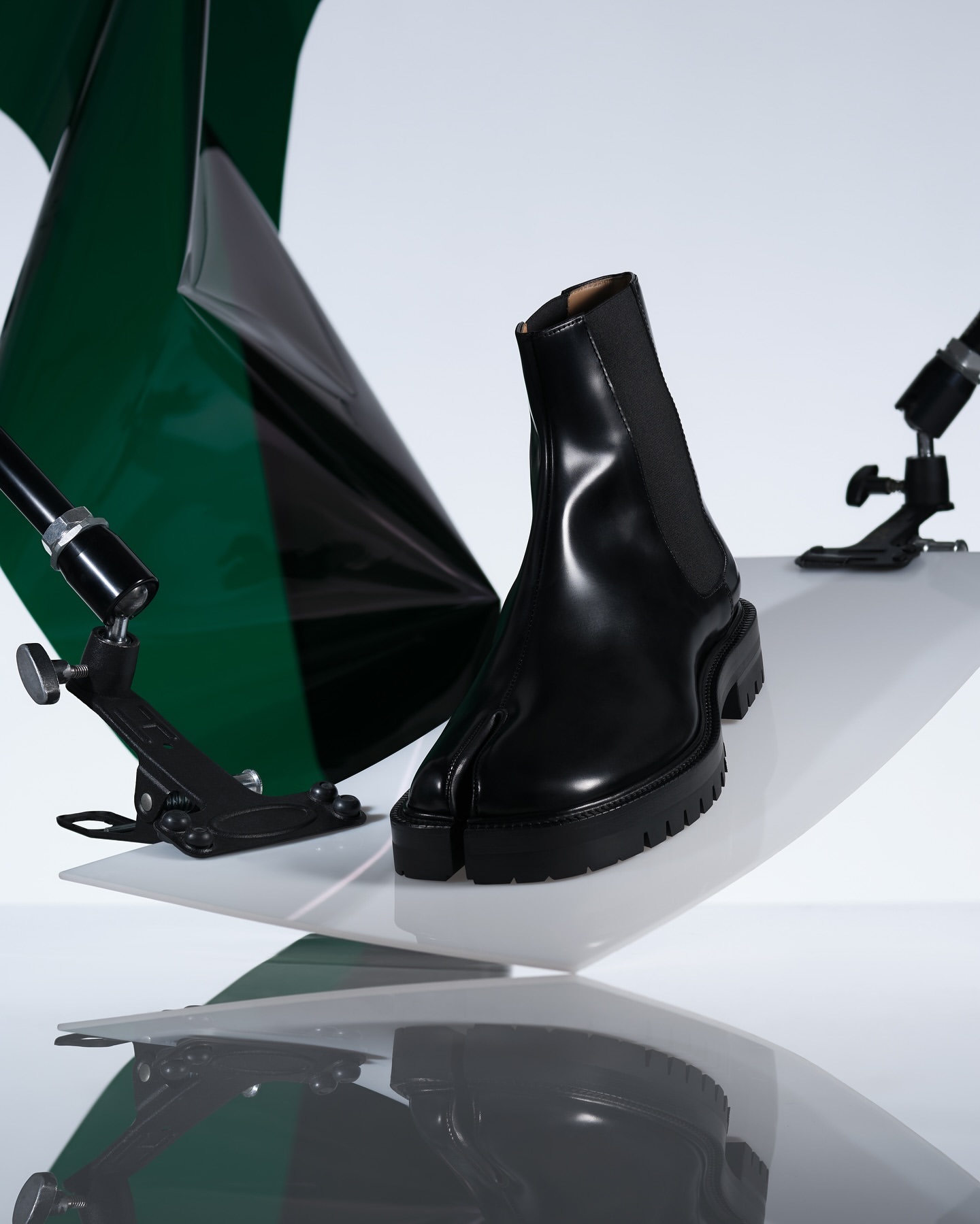 Black leather split-toe ankle boot sits on a white glossy surface with reflective qualities. Green and black shapes in the background.