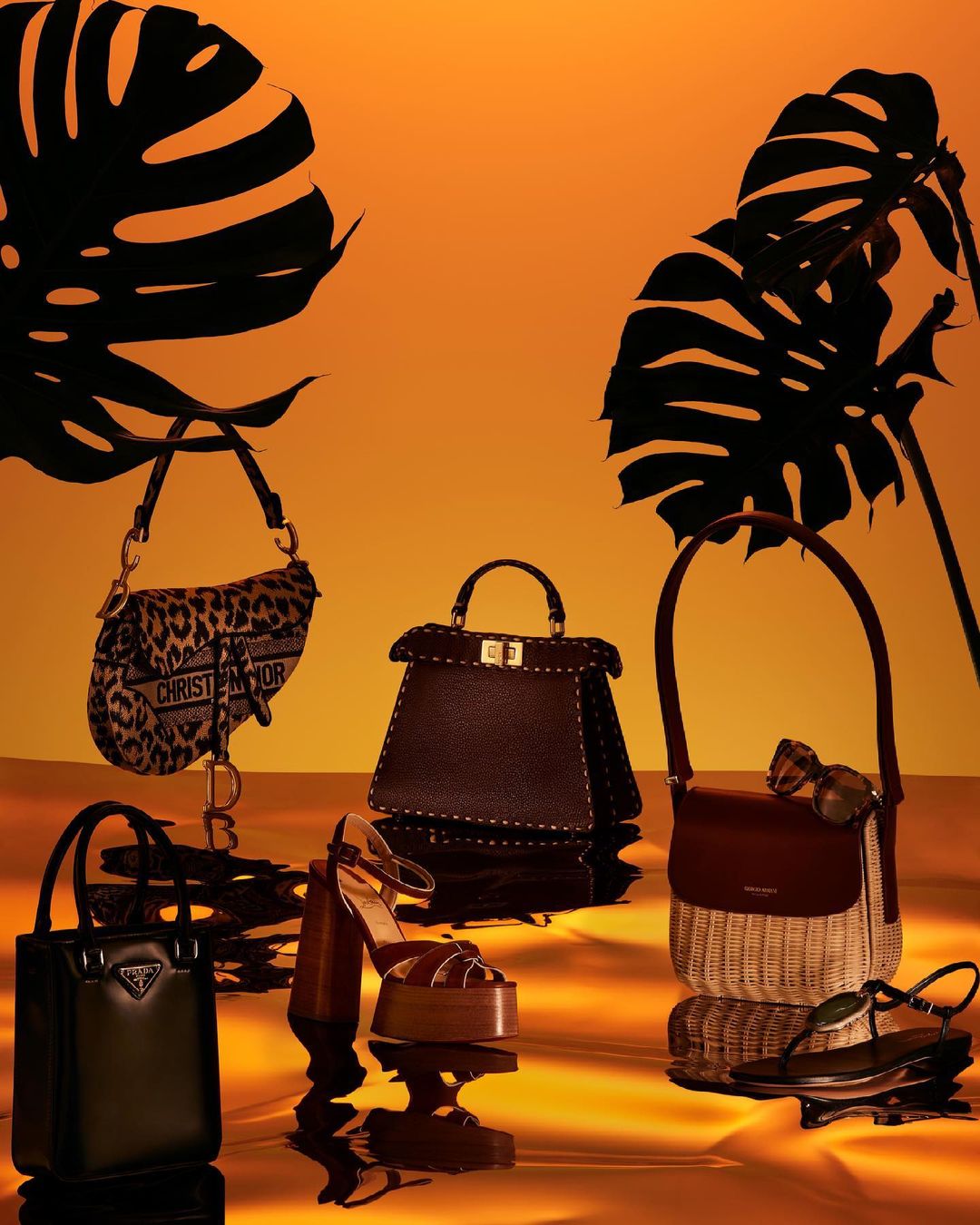 Fashion still life with handbags and sandals on a reflective surface with silhouetted monstera leaves against an orange background.