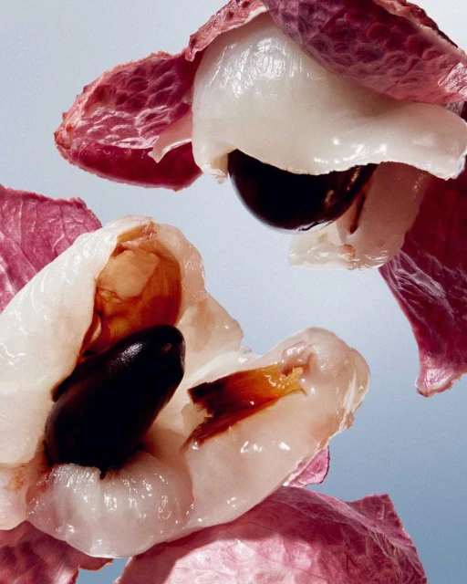 Macro shot of lychee fruits with dark seeds, light flesh, and pink outer skin, set against a pale blue background in studio lighting.