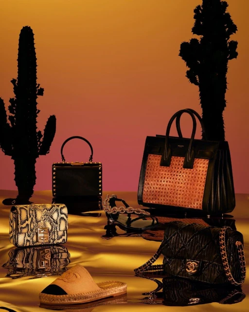 Luxury fashion still life featuring designer handbags, shoes and accessories on a glossy surface, with a desert-themed background.