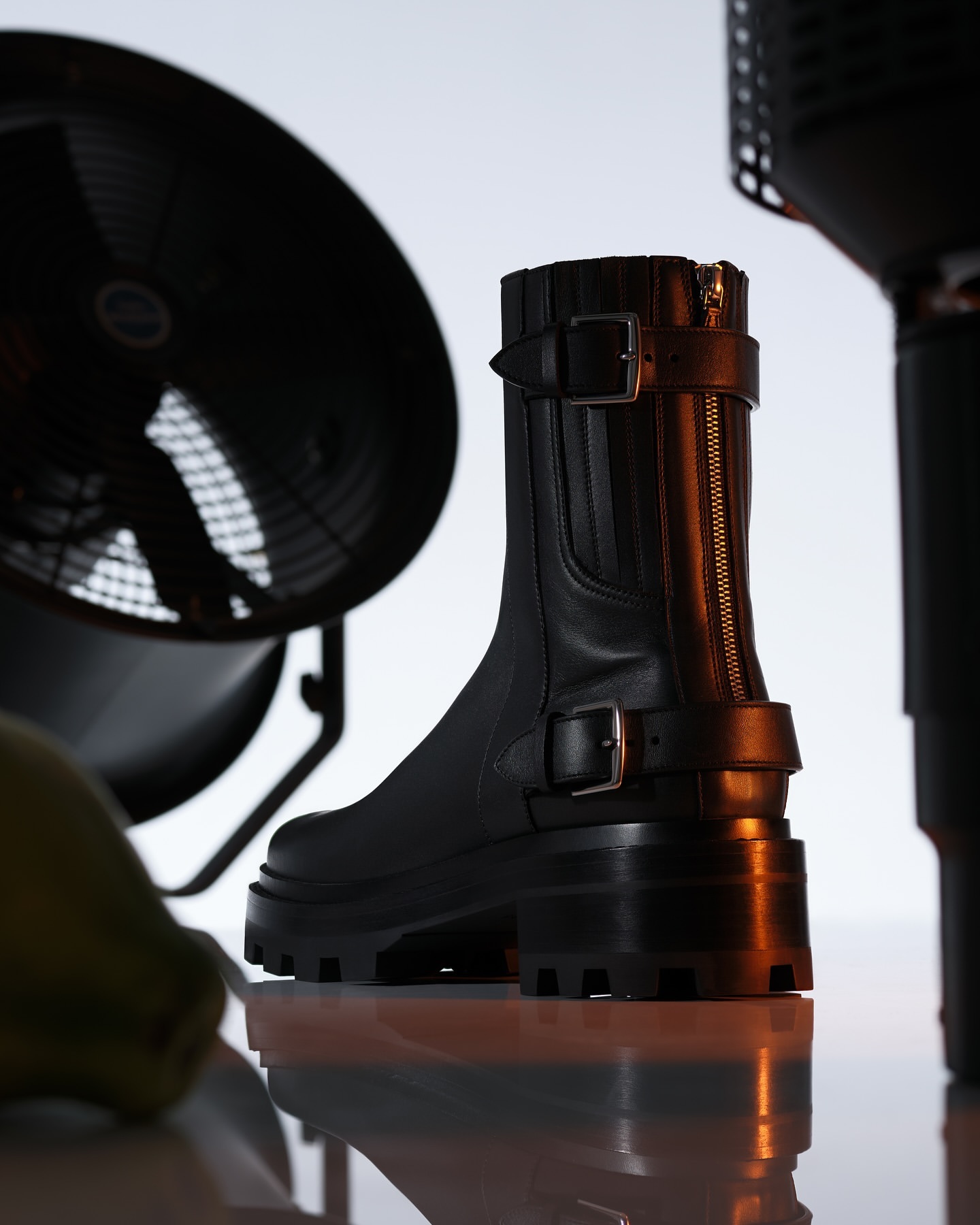 Black leather ankle boot with silver buckles, zippers, and chunky lug sole, positioned in front of black industrial fans.