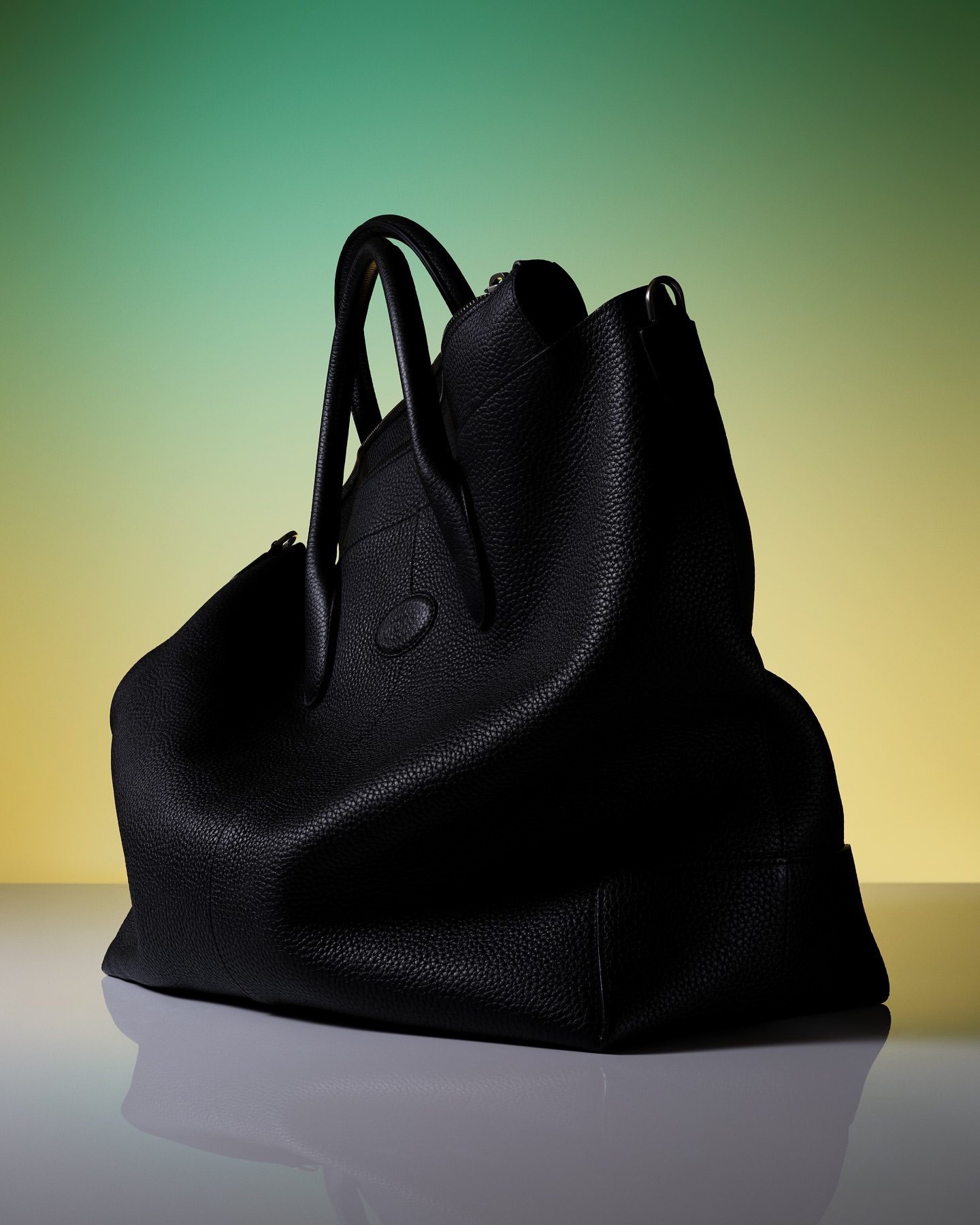 Studio shot of a black pebbled leather handbag with rolled handles against a green and yellow graduated backdrop. Fashion accessory.