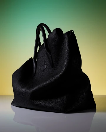 Studio shot of a black pebbled leather handbag with rolled handles against a green and yellow graduated backdrop. Fashion accessory.