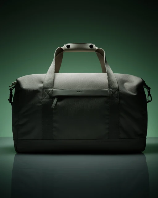 Studio shot of a gray duffel bag featuring a textured design, handle, and front pocket, set against a green backdrop with studio lighting.