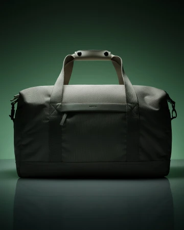 Studio shot of a gray duffel bag featuring a textured design, handle, and front pocket, set against a green backdrop with studio lighting.