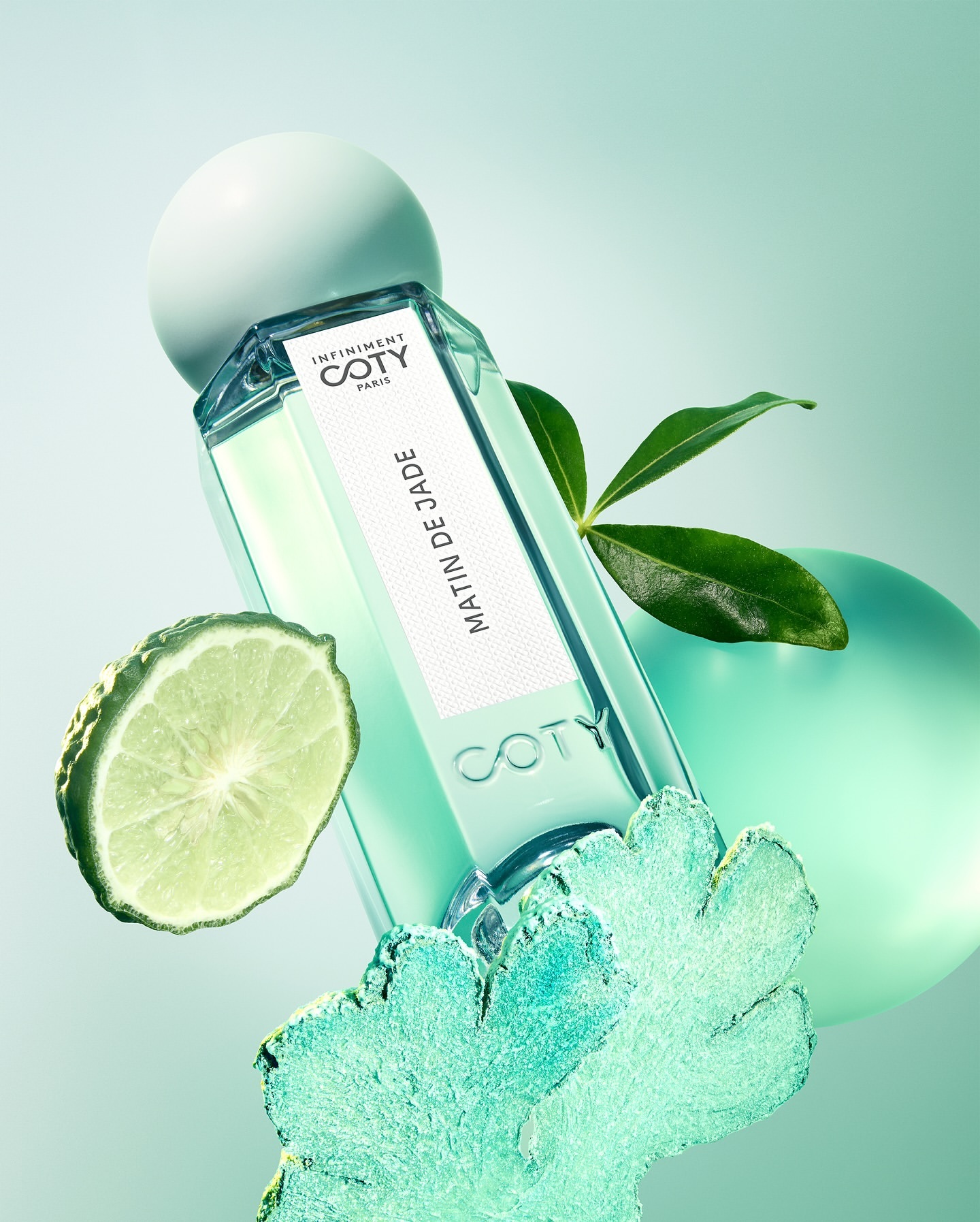 Coty Matin de Jade fragrance: clear glass bottle with green-tinted liquid, styled with a bergamot slice, leaves, and decorative spheres.