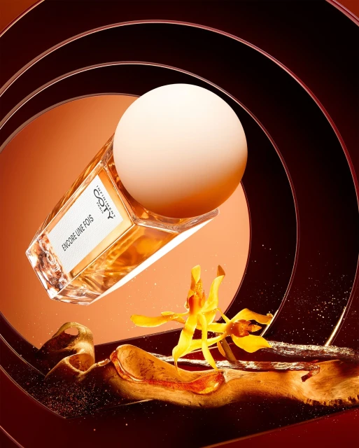 Still life shot of a Coty perfume bottle with a spherical cap, an orchid, and brown resin swirls against a spiral backdrop.