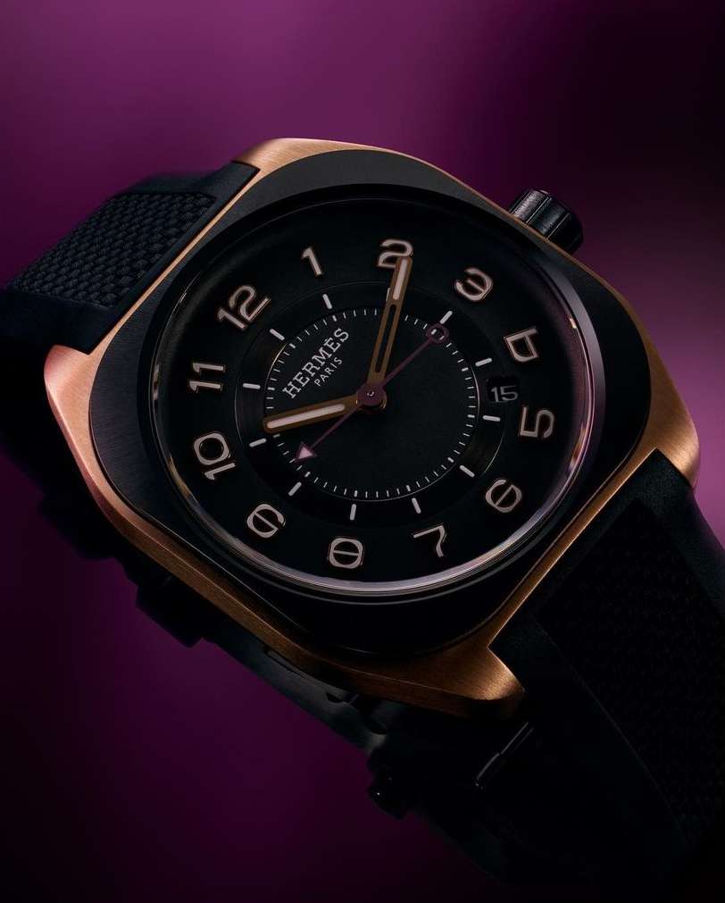 Close-up of a bronze and black Hermès watch with a black leather band, set against a solid purple background, showcasing the brand's craftsmanship.