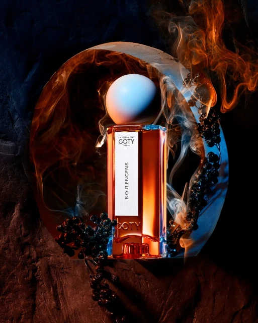 Coty Noir Encens fragrance bottle with spherical cap, surrounded by smoke and berries on a dark rock surface. Studio product shot.