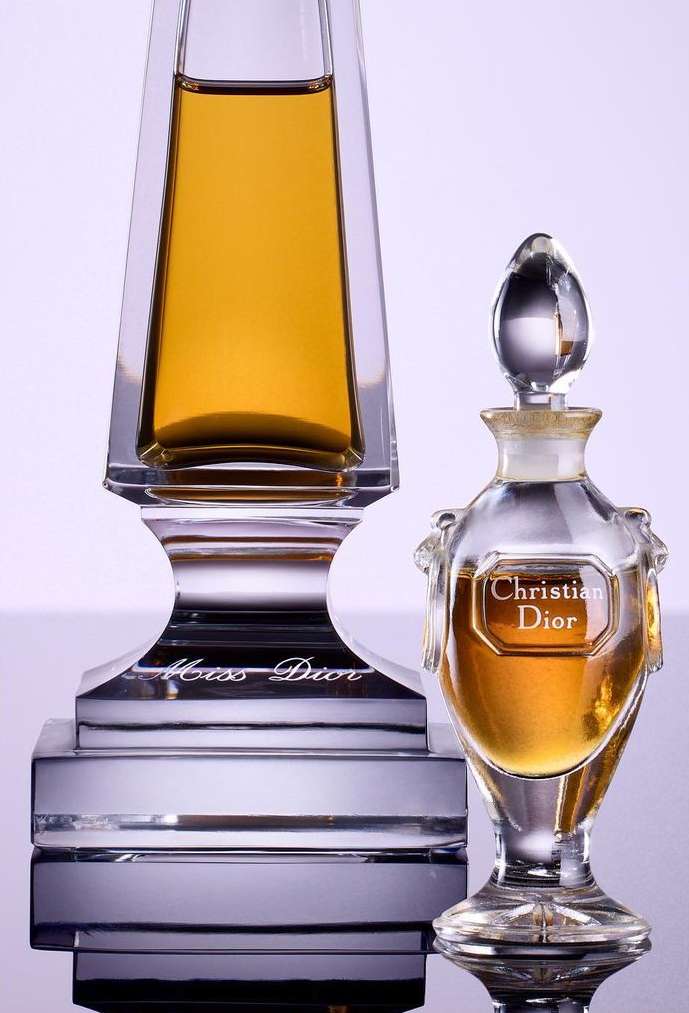 Two glass perfume bottles, one taller and one shorter, both filled with brown liquid, are on display in a studio setting with soft lighting.