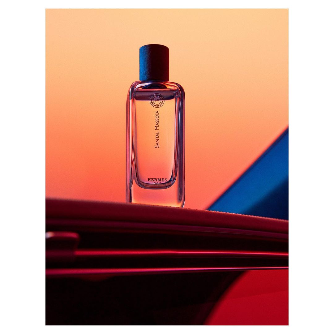 Studio shot of a clear glass Hermes Santal Massoïa perfume bottle, featuring an orange and blue backdrop and a dark-red reflective surface.