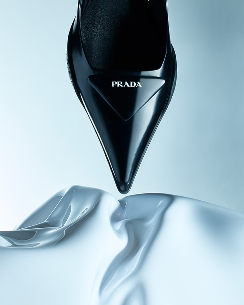 Close-up studio shot of a glossy black Prada stiletto heel shoe resting on a flowing white silk cloth, set against a light blue background.