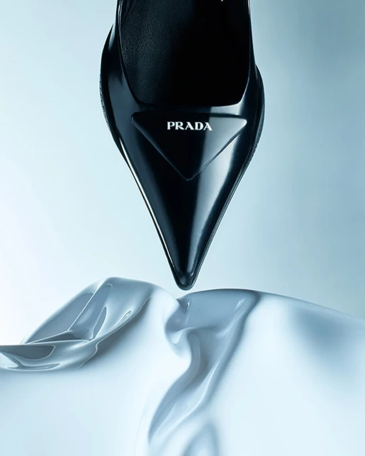 Close-up studio shot of a glossy black Prada stiletto heel shoe resting on a flowing white silk cloth, set against a light blue background.