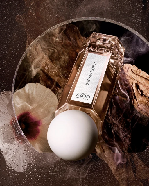 Still life shot of a golden Coty perfume bottle lying horizontally, with a white sphere, flower, and wood against a dark backdrop.
