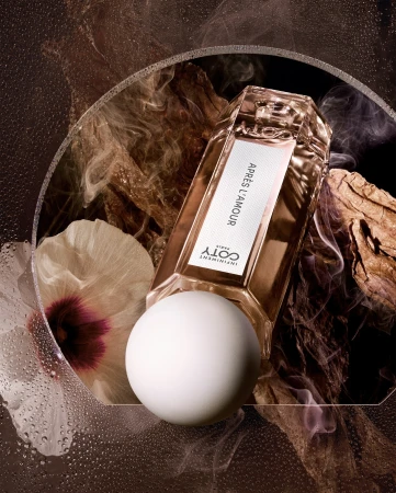 Still life shot of a golden Coty perfume bottle lying horizontally, with a white sphere, flower, and wood against a dark backdrop.