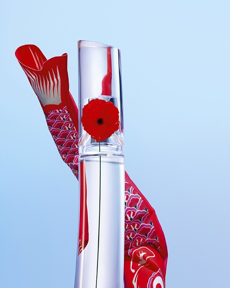 Still life of a clear glass bottle with a red flower inside, next to a red and white fish-shaped decoration on a gradient blue background.