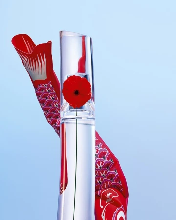 Still life of a clear glass bottle with a red flower inside, next to a red and white fish-shaped decoration on a gradient blue background.