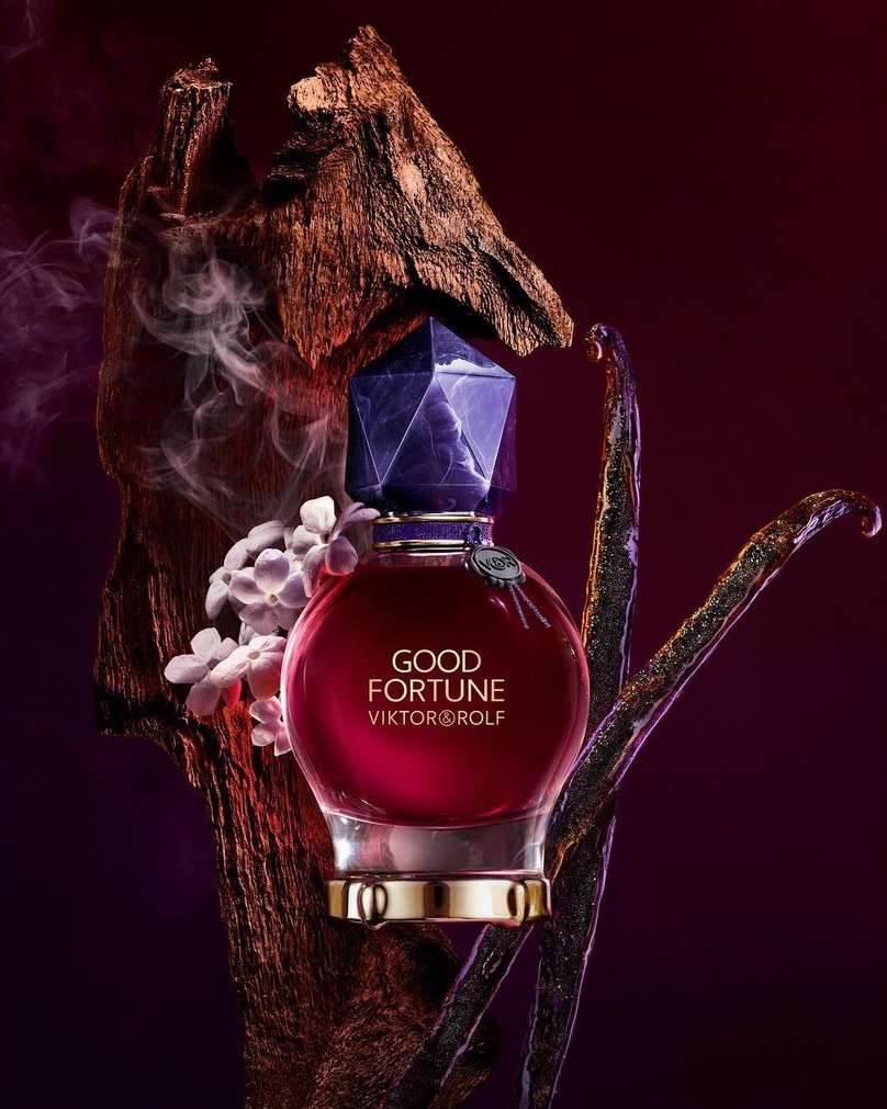 Close-up product shot of Good Fortune perfume bottle by Viktor&Rolf with a purple cap, surrounded by dried bark, vanilla beans, and white flowers.