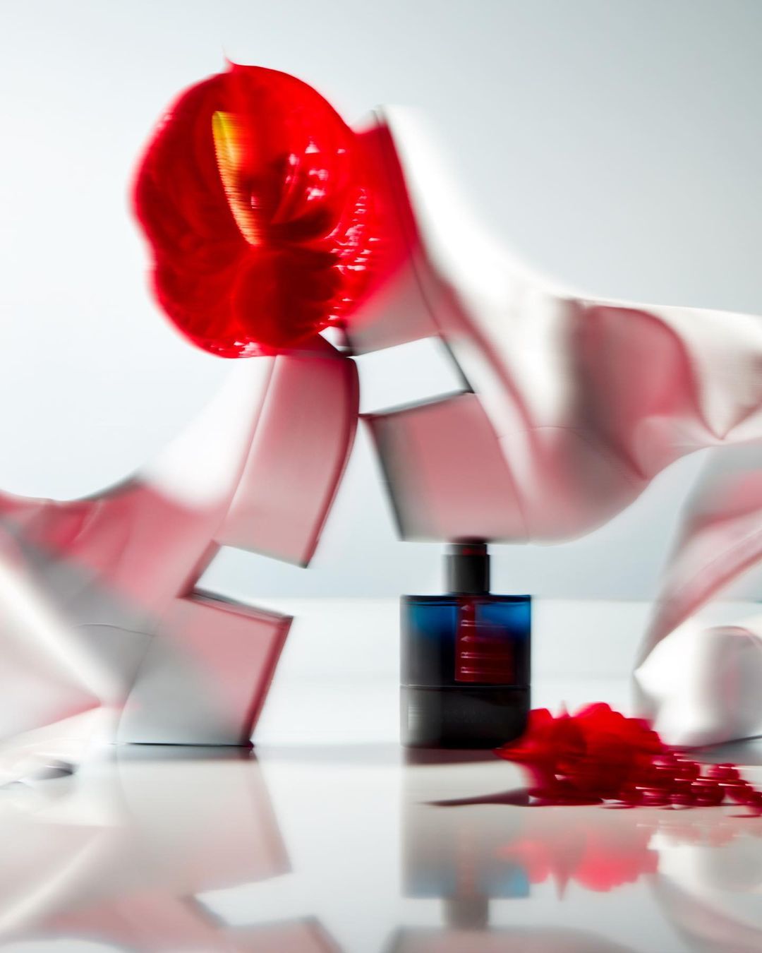 Still life of a perfume bottle, red anthurium flower, and white fabric on a reflective surface in a minimalist, artistic composition.