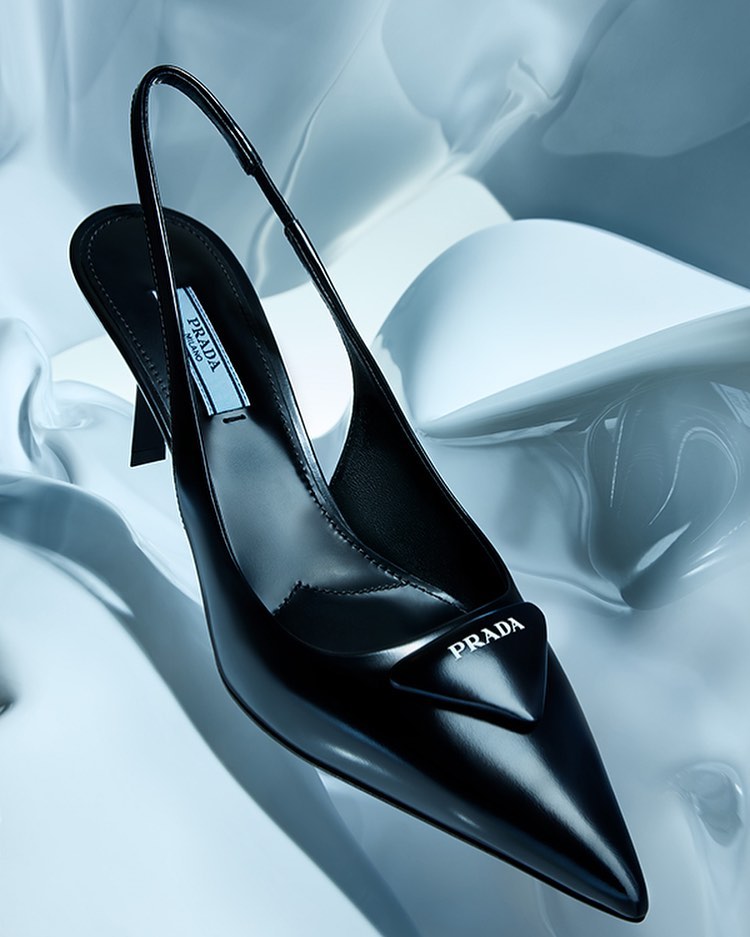 Elegant black slingback pump by Prada on a glossy white surface. High heel, pointed toe, and triangular Prada logo detail.
