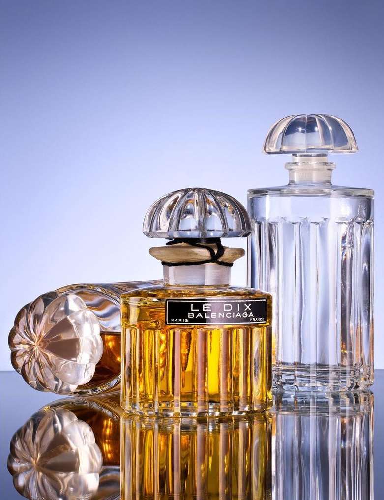 Still life photograph of three vintage Balenciaga perfume bottles with clear glass, ribbed tops, and golden liquid against a gradient blue background.