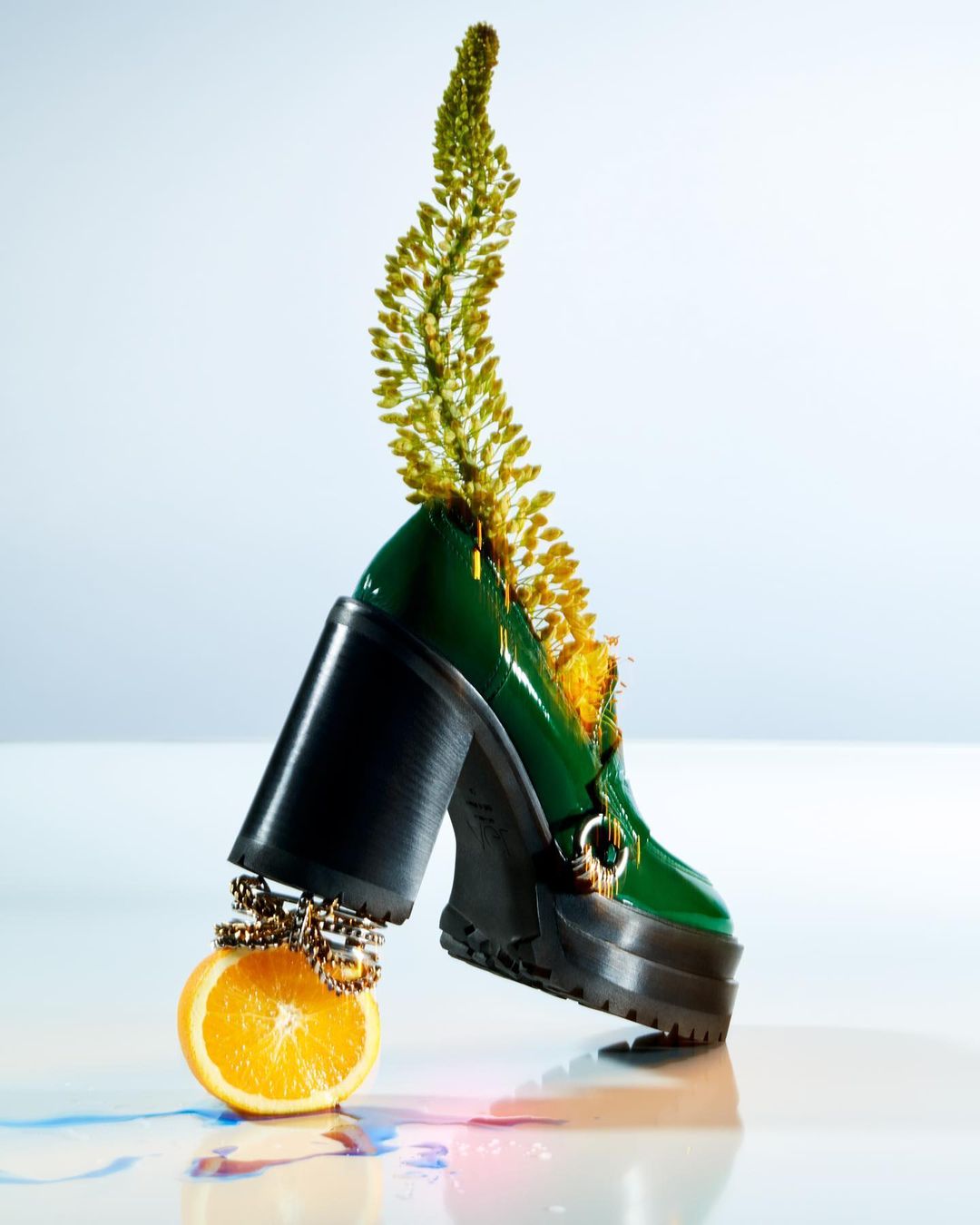 Still life shot of a green heeled loafer with a yellow floral accent balancing on an orange, studio setting.