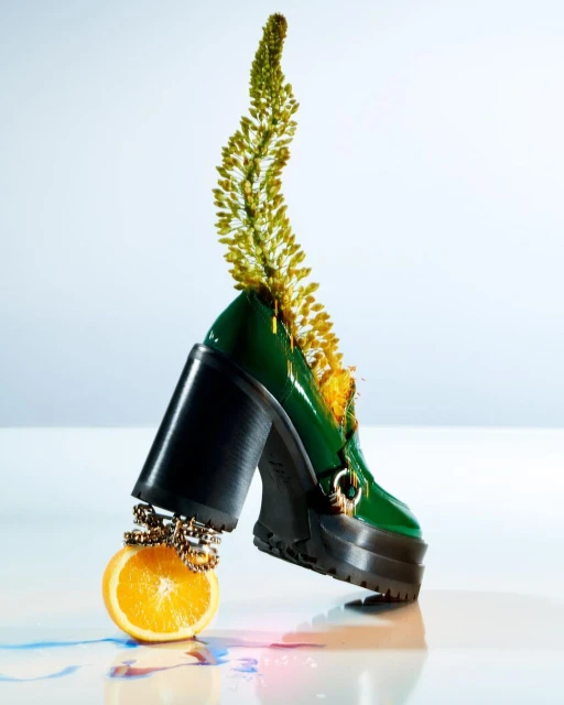 Still life shot of a green heeled loafer with a yellow floral accent balancing on an orange, studio setting.