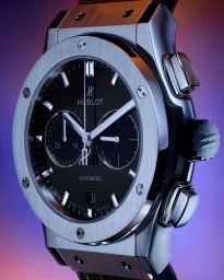 Close-up macro shot of a luxury Hublot chronograph watch with a brushed silver bezel and black dial against a blue and purple gradient background.