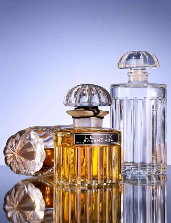 Three crystal perfume bottles with fluted designs and faceted stoppers reflecting on a smooth surface against a soft blue gradient background.