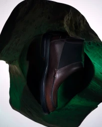 Brown leather Chelsea boot displayed inside a hollow cutout of a textured green stone sculpture against a bright white background.