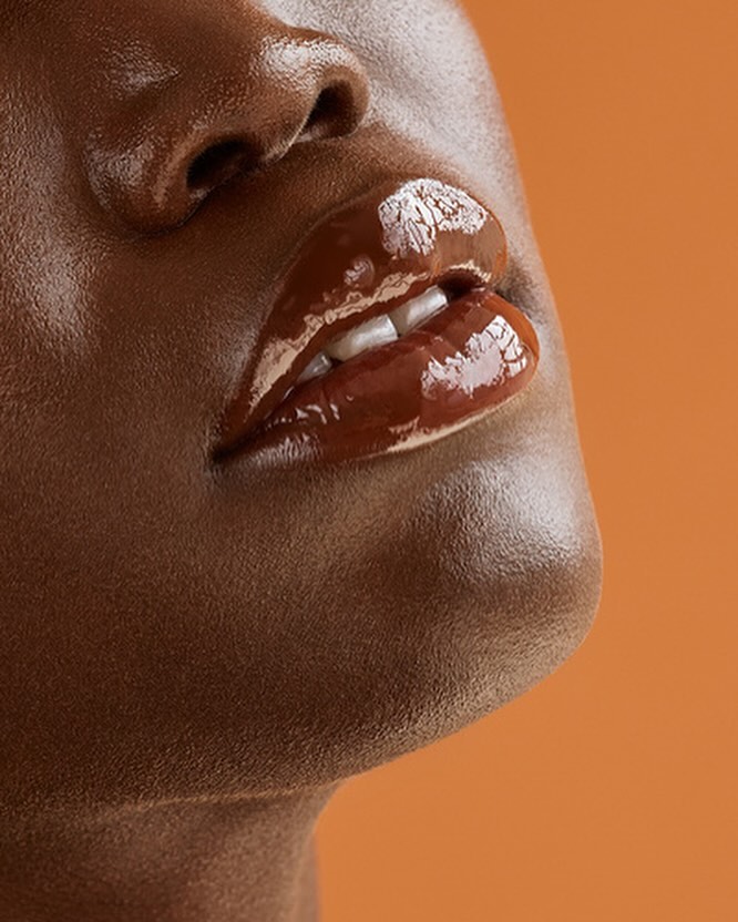 Close-up of an adult female's face featuring dark skin, glossy brown lips, and visible white teeth against a solid orange background.