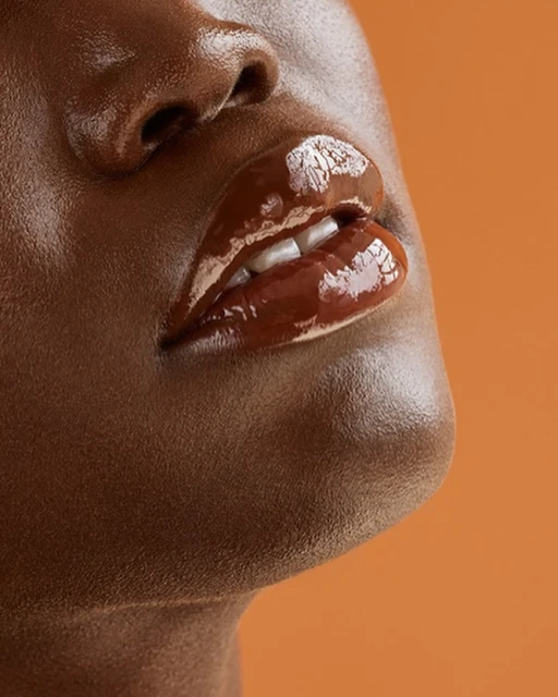 Close-up of an adult female's face featuring dark skin, glossy brown lips, and visible white teeth against a solid orange background.