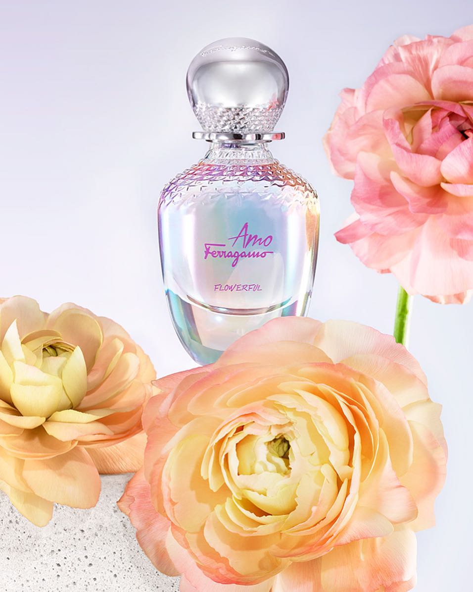Close-up studio shot of a pearlescent perfume bottle with a silver cap, surrounded by peach-colored ranunculus flowers against a white background.