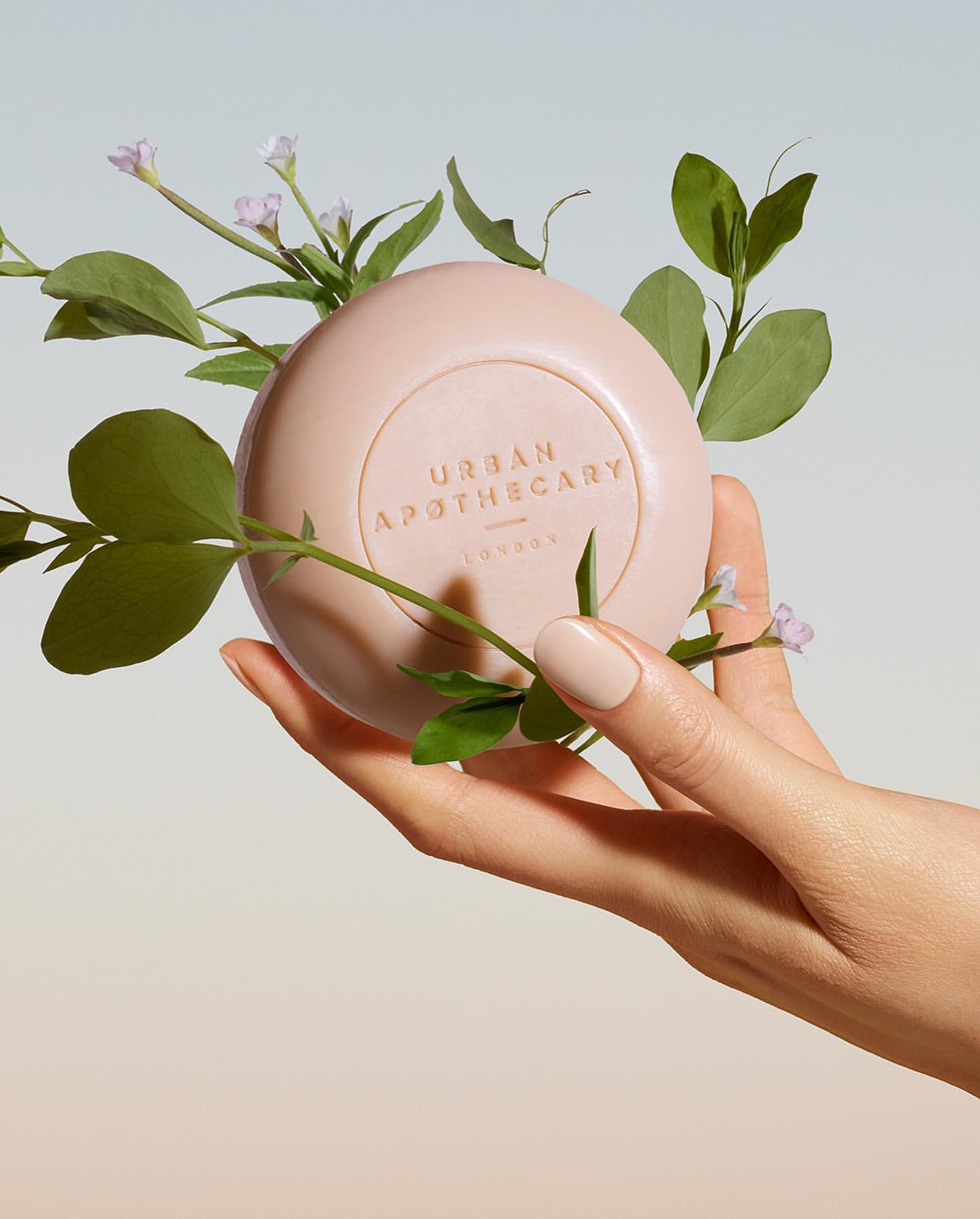 Woman's hand holding a round, light pink soap surrounded by green leaves and small purple flowers, product shot for beauty and wellness.