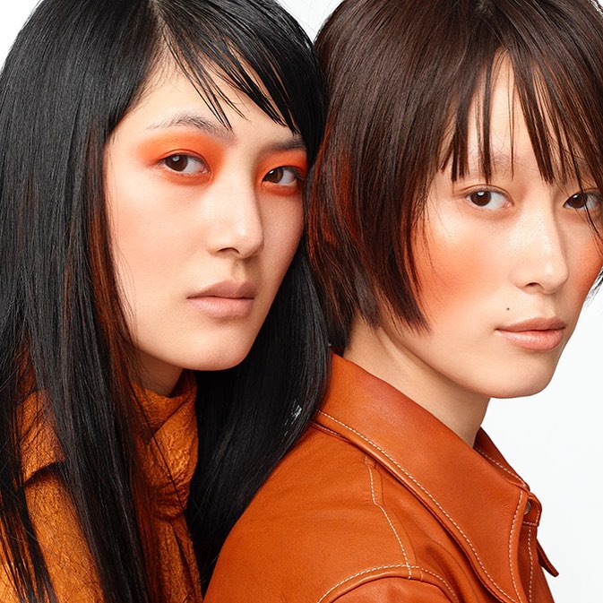 Two Asian women pose closely in a beauty shot. They wear orange eye shadow and matching outfits. One has black hair and the other brown.