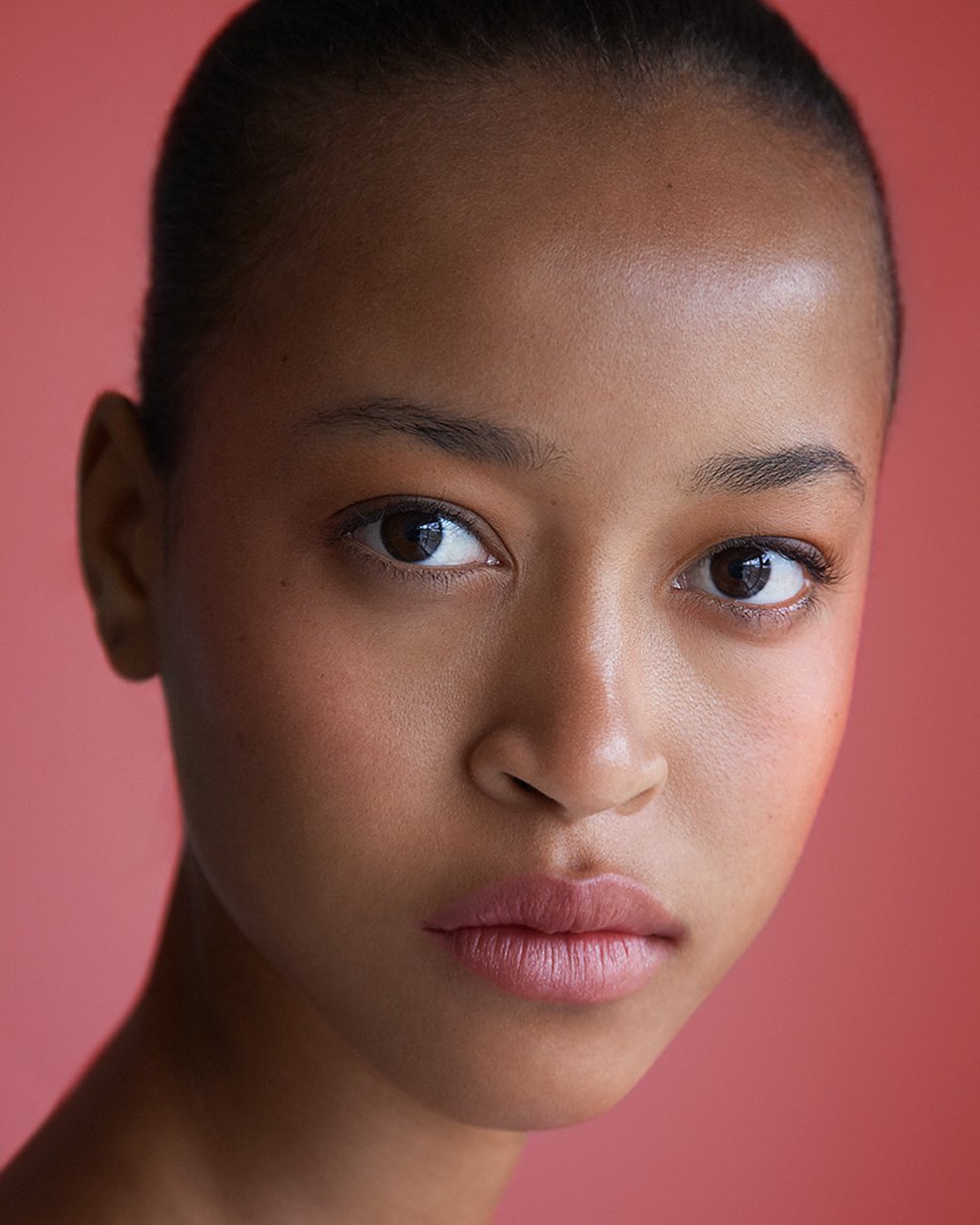 Close-up portrait of a young adult woman with brown eyes and light pink lips against a blurred, coral-colored background, showcasing natural skin.