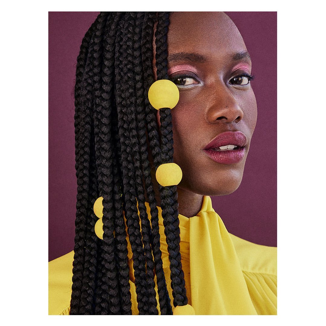 Close-up of a Black woman with braided hair adorned with yellow balls, wearing a yellow top against a purple background.