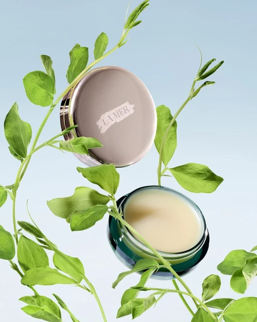 La Mer skincare product display with face cream in a jar and lid, accented by green leafy plant stems against a gradient blue backdrop.