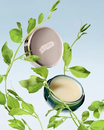 La Mer skincare product display with face cream in a jar and lid, accented by green leafy plant stems against a gradient blue backdrop.