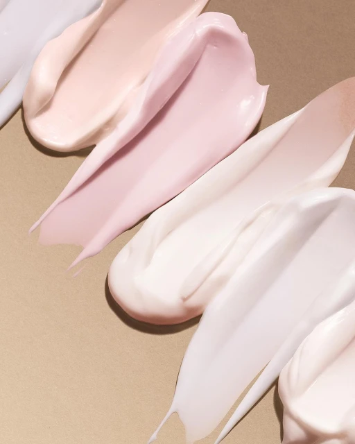 Cosmetic product swatches in varying shades of cream and pink are displayed on a tan surface, showcasing texture and color.