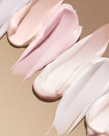 Cosmetic product swatches in varying shades of cream and pink are displayed on a tan surface, showcasing texture and color.