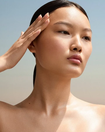 Close-up portrait of an East Asian woman with glowing skin touching her forehead against a clear sky background
