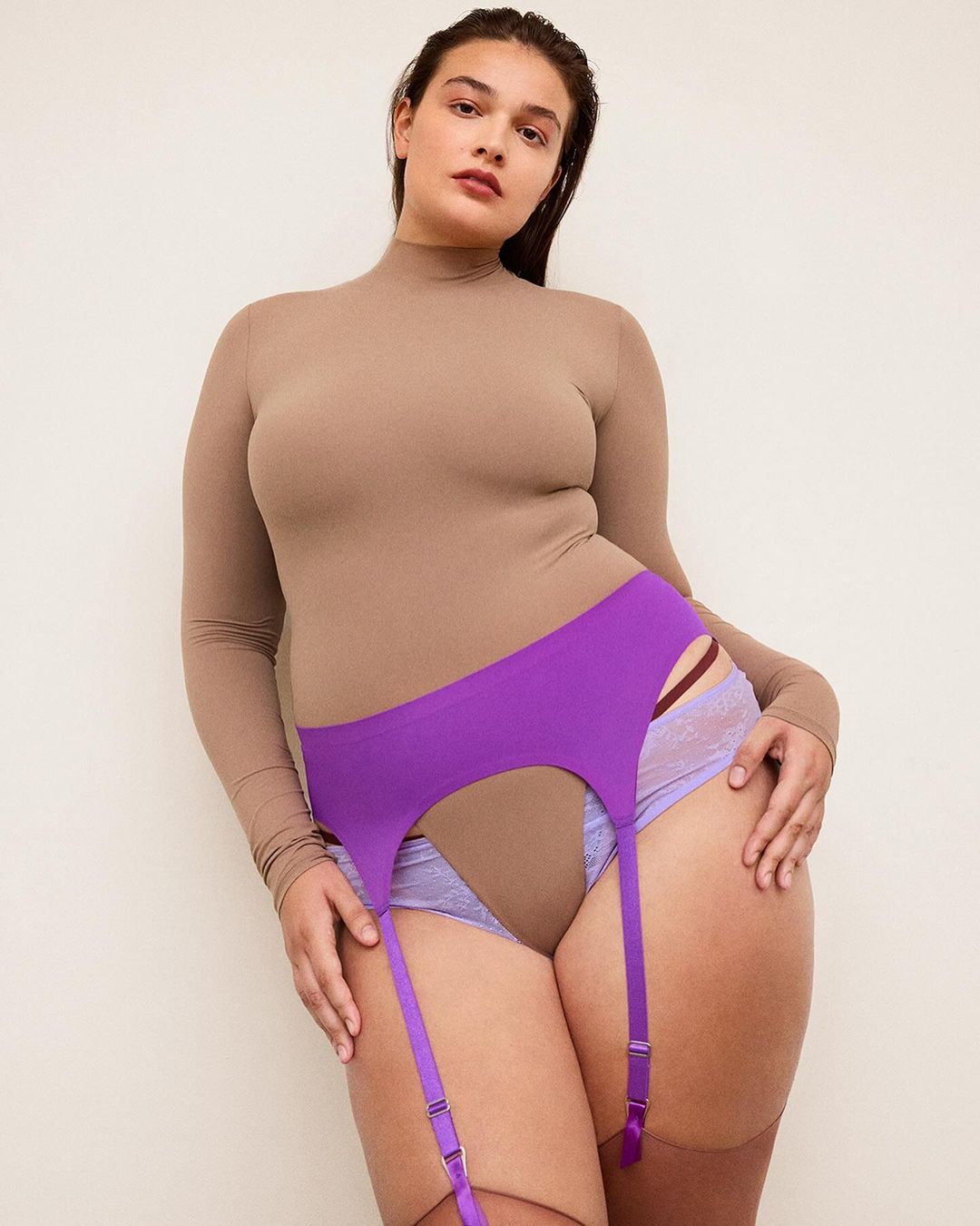 Studio shot of a woman in a bodysuit and garter belt, posing against a white backdrop. The focus is on fashion and body positivity.
