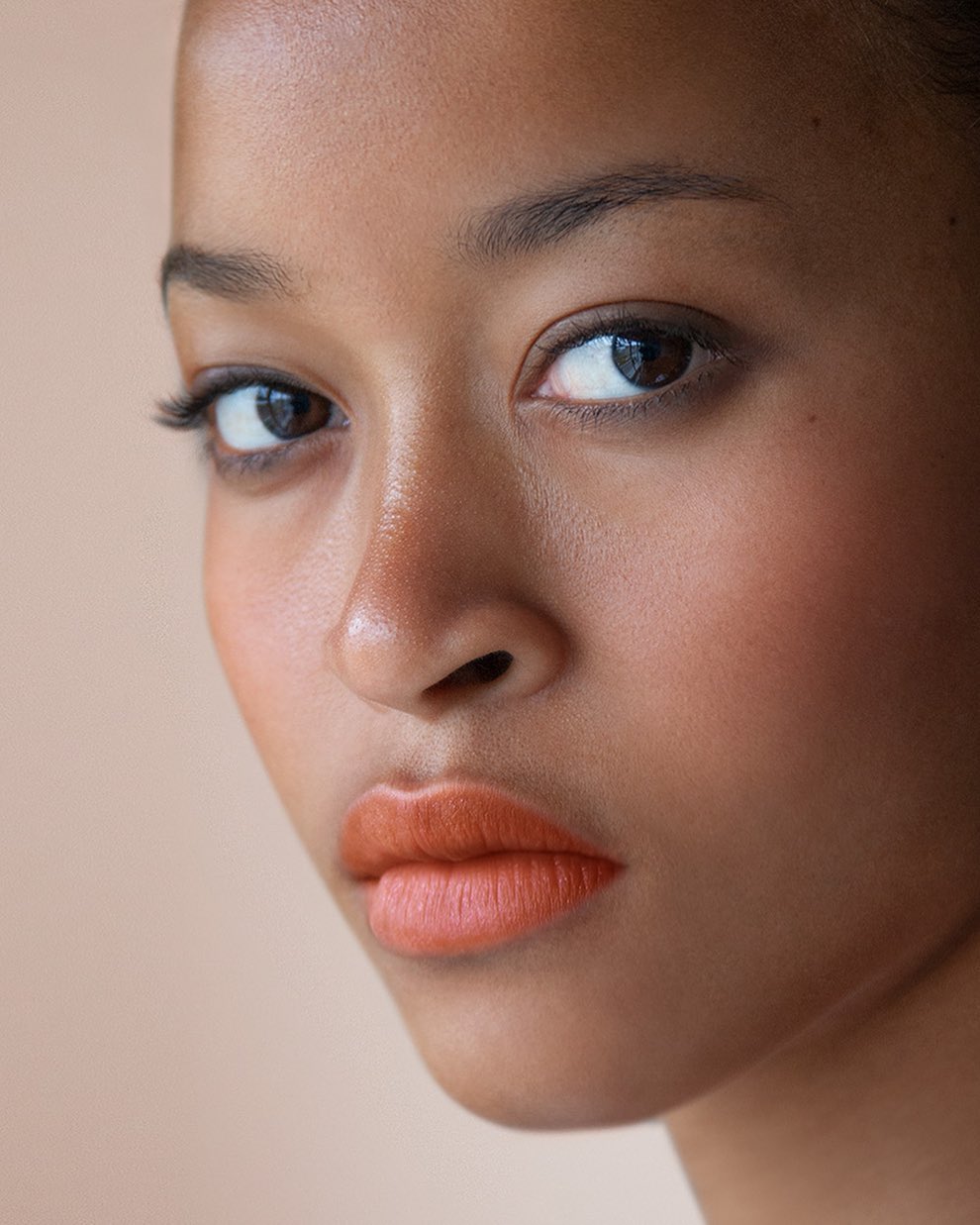 Close-up portrait of a young adult woman with dark hair and brown eyes, wearing coral lipstick against a neutral beige background.
