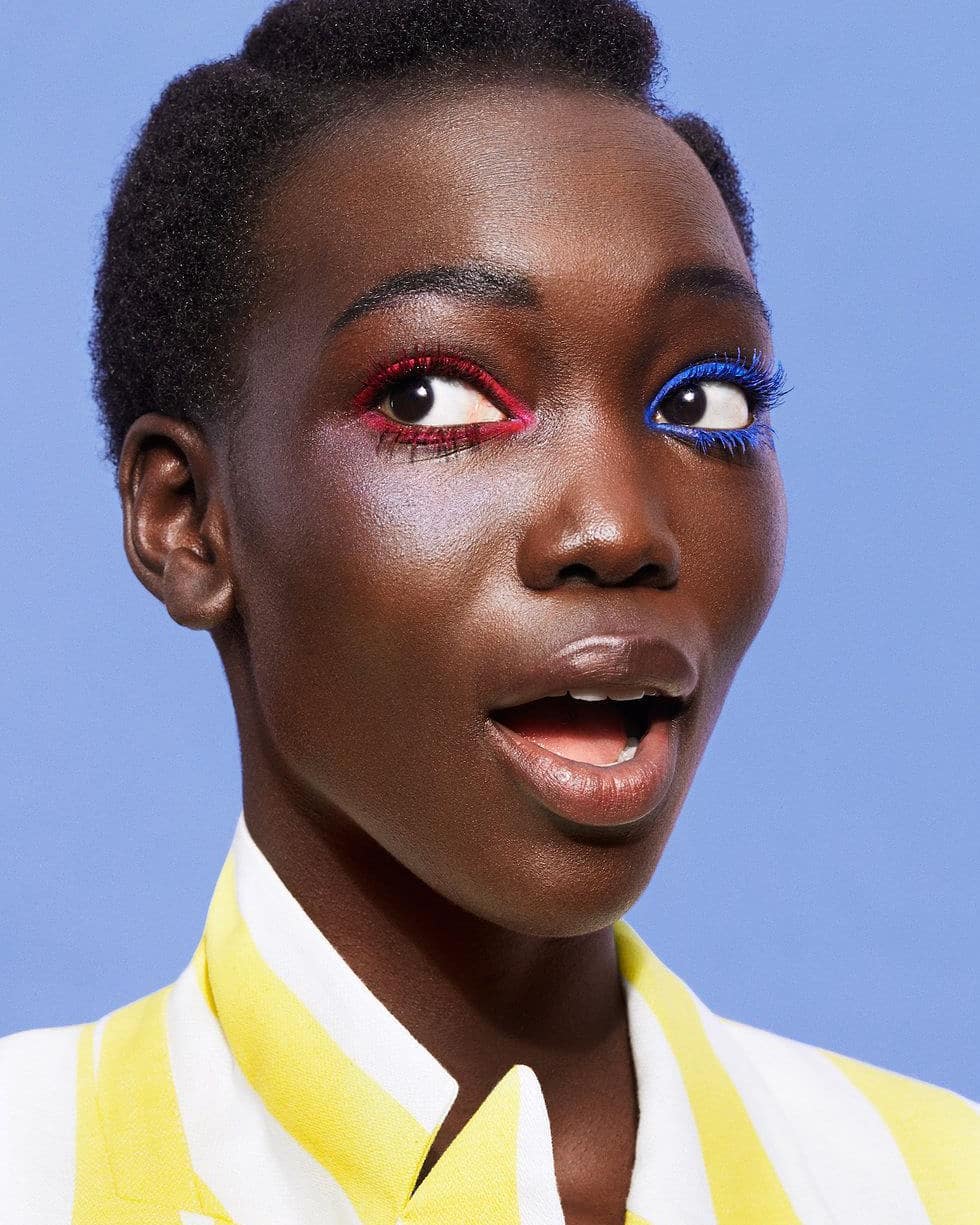 Close-up beauty shot of a dark-skinned Black woman with colorful makeup wearing a yellow and white striped jacket against a blue backdrop.