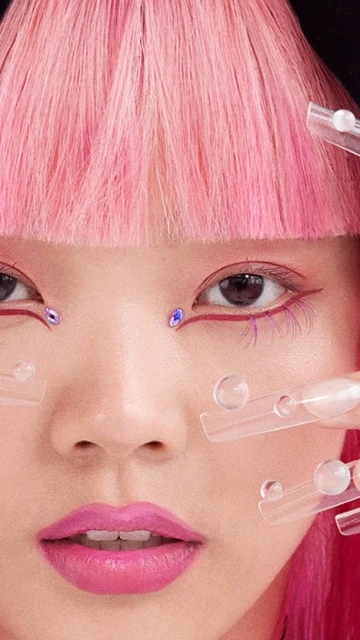 Close-up of a woman with pink hair and creative makeup, including gems around her eyes and clear acrylic nail art on her face.