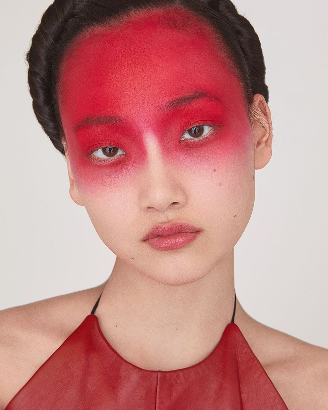 Portrait of a young woman with red face paint and a braided hairstyle, wearing a red leather halter top against a neutral background.